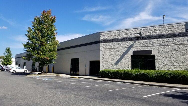 Exterior of a single-story stone-faced warehouse and office building with a parking lot and trees under a blue sky.