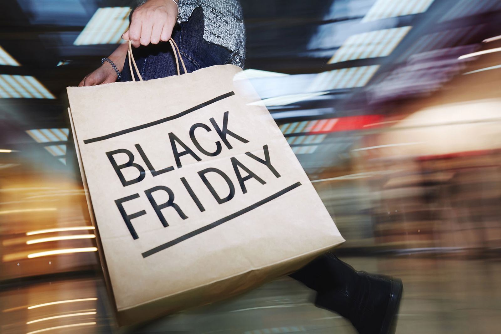Person holds a large brown shopping bag with “BLACK FRIDAY” text, walking fast in a blurred, colorful mall setting, creating motion.