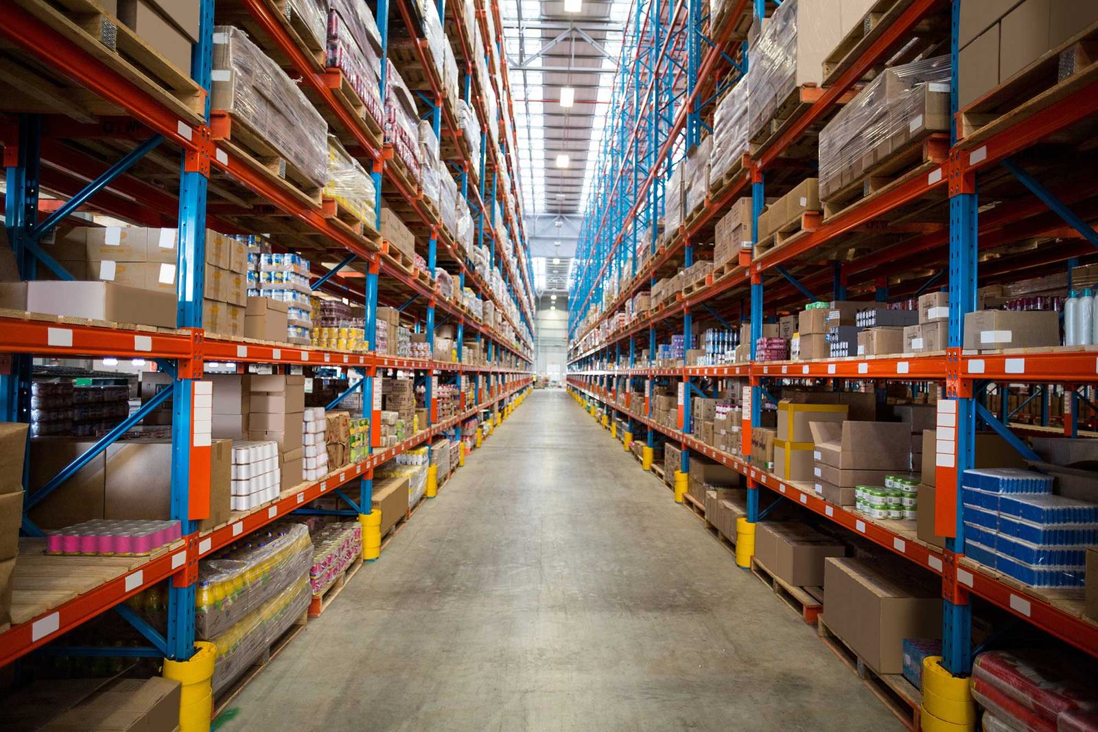 Warehouse interior with tall shelves packed with boxes and goods. The aisle is clear, and the setting is brightly lit, orderly, and spacious.