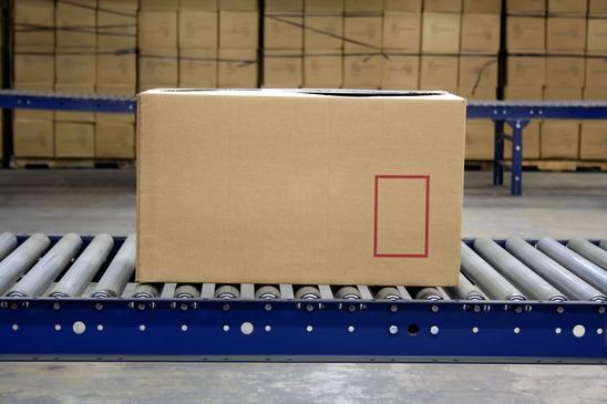 Cardboard box on a roller conveyor in a warehouse, with stacked boxes in the background.