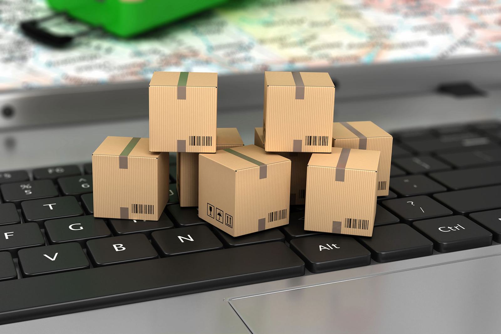 Small cardboard boxes on a laptop keyboard symbolize e-commerce. Blurred map and green item in the background. Boxes have barcodes.