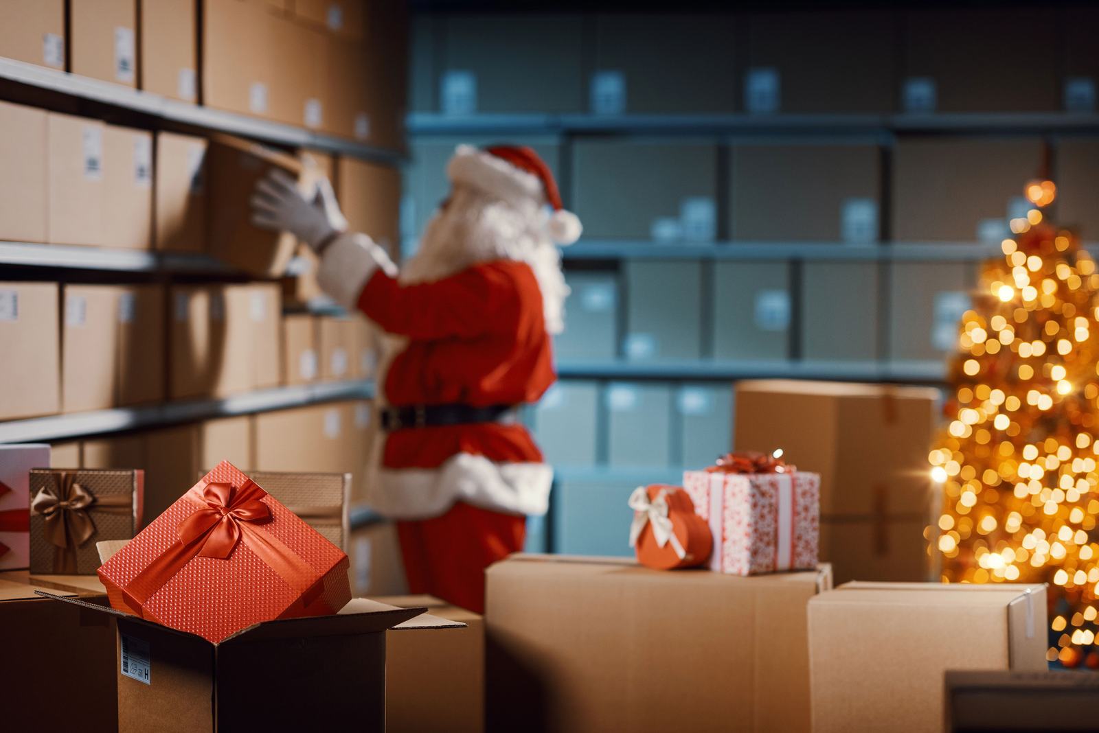 Santa in a storeroom stacks boxes. A glowing Christmas tree stands nearby. Gift boxes with red ribbons are in the foreground. Festive mood.